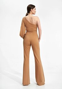 Een bruin jumpsuit met één schouder, een nauwsluitende lijfje en flared broekspijpen, gemaakt van een gladde stof, met een poffende mouw aan één kant.