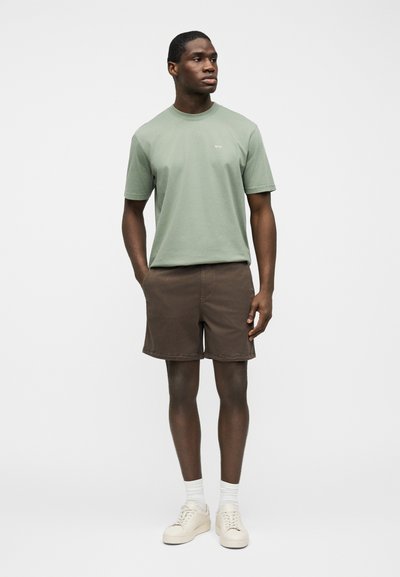 Homme portant un t-shirt vert sauge à manches courtes, un short marron, des chaussettes blanches et des baskets crème, debout devant un fond blanc uni.