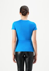 Vrouw met haar in een knot, gekleed in een felblauw shirt met korte mouwen en zwarte leren broek, staande tegen een witte achtergrond.