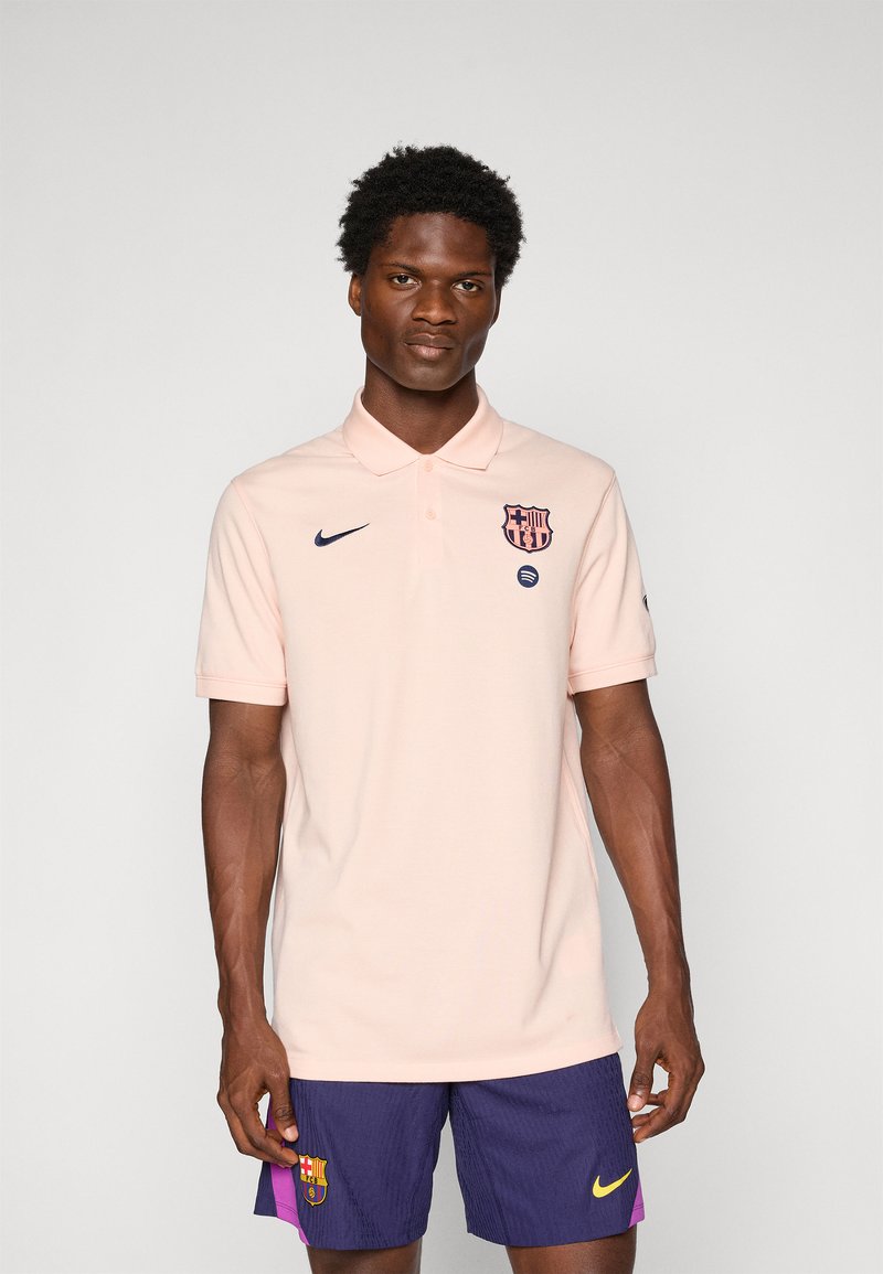 Man wearing light pink polo with FC Barcelona and Nike logos, paired with navy blue FC Barcelona shorts with a yellow Nike swoosh.