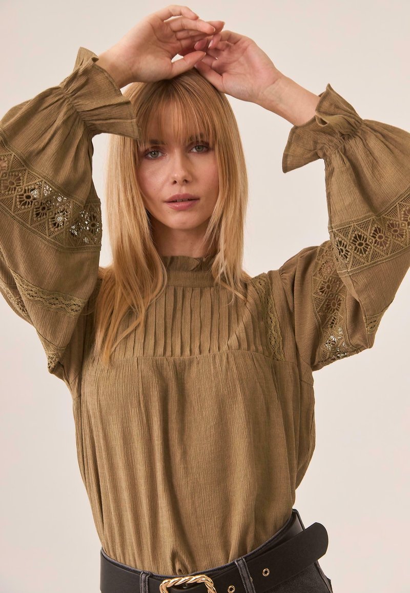 Mujer con una blusa marrón suelta con detalles de encaje y mangas anchas, levantando ambas manos por encima de su cabeza frente a un fondo neutro.