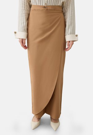 Donna che indossa una gonna a portafoglio a vita alta color cammello sopra pantaloni abbinati, con décolleté beige con punta appuntita e camicia bianca a righe con bottoni ai polsini.
