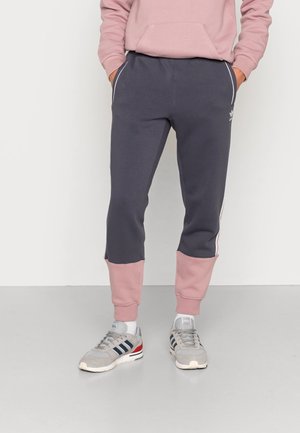 Person trägt dunkelgraue Jogginghose mit pinken Bündchen und Seitenstreifen, pinken Hoodie und graue Sneakers, steht mit Händen in den Taschen.