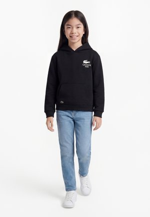 Ragazza che cammina in avanti indossando una felpa nera Lacoste con cappuccio, jeans azzurri chiari e sneakers bianche su uno sfondo bianco semplice.