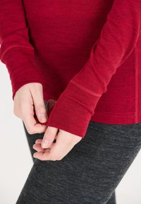 Langarmige rode top gemaakt van zachte stof, met duimgaten en contrasterende naden, gecombineerd met donkergrijze leggings.