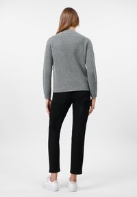 Woman with long brown hair wearing a grey knit sweater, black pants, and white sneakers, standing facing away against a white background.