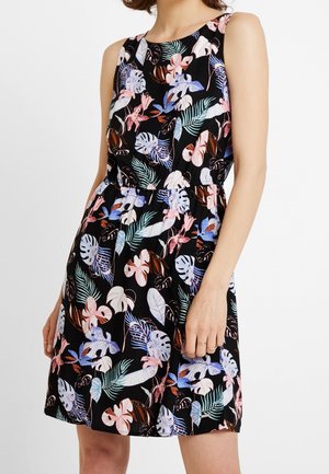 Czarna sukienka bez rękawów z różowymi, niebieskimi i białymi tropikalnymi kwiatowymi i liściastymi wzorami, dopasowana w talii, noszona przez stojącą osobę.