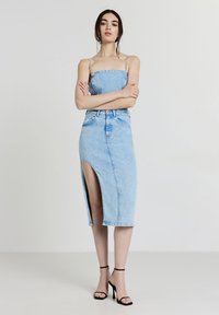 Femme portant un top sans manches en denim bleu clair et une jupe midi assortie avec fente à l'avant, assortie à des sandales à talons hauts noires, se tenant les bras croisés.