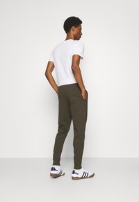 Weißes T-Shirt mit kurzem Arm, dunkelgrüne Jogginghose mit Seitentaschen und weiße Sneakers mit marineblauen Akzenten. Einfarbiger Hintergrund.