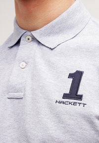 Detailní záběr na světle šedý límeček polokošile se dvěma knoflíky, s námořnickou vyšitou číslicí "1" a textem "Hackett" na hrudi.