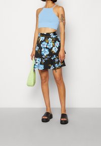 Light blue cropped tank top, black floral skirt with blue and green flowers, green woven bag, black platform sandals. Smooth texture throughout.
