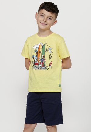 T-shirt jaune avec un motif de planche de surf et des palmiers. Le tissu semble léger. Associé à un short bleu marine foncé avec une pièce visible sur la jambe.