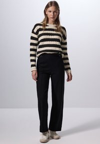 Schwarzer und weißer gestreifter Strickpullover mit geripptem Muster, kombiniert mit hochgeschnittenen schwarzen Hosen, die seitliche Knöpfe und einen maßgeschneiderten Schnitt aufweisen.