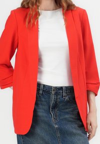 Czerwony żakiet o dopasowanym kroju, z rękawami o długości trzech czwartej i gładką fakturą, zestawiony z białą bluzką oraz niebieską spódnicą dżinsową.