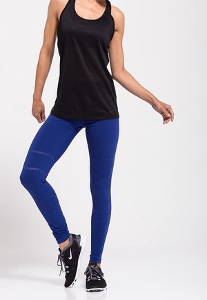Femme portant un haut noir sans manches, des leggings bleus et des chaussures de course Nike noires, debout avec un pied légèrement levé.
