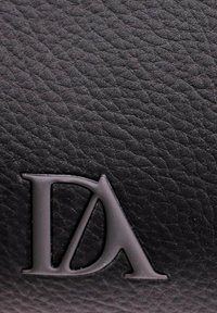 Superficie de cuero negro texturizado con emblema en relieve mate negro "DA" en la esquina inferior izquierda.