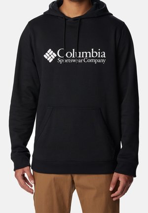 Homme portant un sweat à capuche noir Columbia Sportswear Company avec poche avant et pantalon beige, montré du cou aux hanches sur fond blanc.
