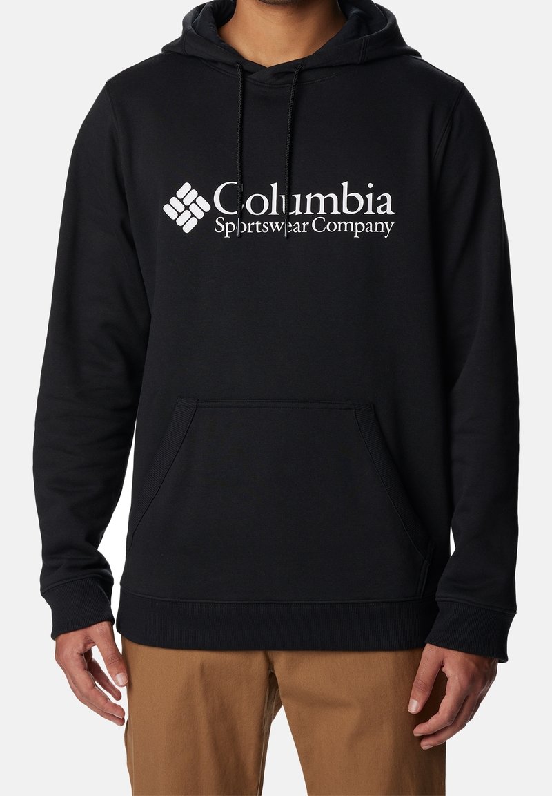 Homme portant un sweat à capuche noir Columbia Sportswear Company avec poche avant et pantalon beige, montré du cou aux hanches sur fond blanc.