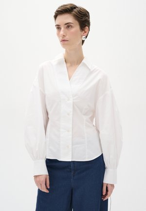 Blusa di cotone bianca con scollo a V, chiusura con bottoni e maniche a sbuffo. Indossata con pantaloni in denim blu scuro. Texture liscia, vestibilità tailorata.