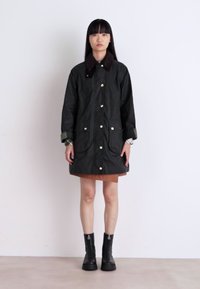 Dark green waterproof jacket with a brown corduroy collar, gold snap buttons, and front pockets. Worn over a brown skirt and black boots.