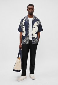 Λ navy κοντομάνικο πουκάμισο με άσπρα λουλούδια και αστέρια, συνδυασμένο με μαύρο παντελόνι και άσπρα αθλητικά παπούτσια. Κρατάει μια μπεζ τσάντα με ναυτικές λεπτομέρειες.