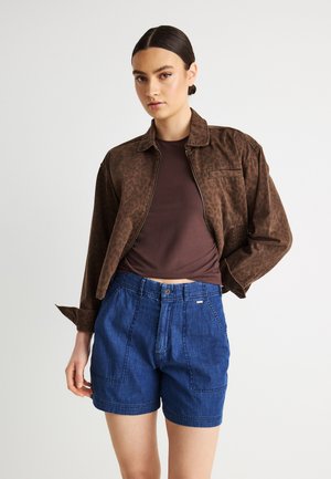 Frau mit dunklem, zum Dutt gebundenem Haar, die eine braun gemusterte Jacke, ein dunkelbraunes Oberteil und hochtaillierte blaue Jeansshorts vor weißem Hintergrund trägt.