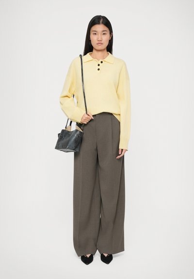 Donna in un maglione giallo a maniche lunghe e pantaloni grigi a gamba larga con una borsa nera appesa alla spalla contro uno sfondo uniforme.