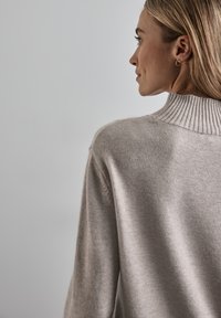 Světle béžový pletený svetr s vysokým vroubkovaným límcem, uvolněným střihem a měkkou texturou, zobrazený z bočního a zadního úhlu.