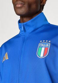 Μπλε αθλητικό μπουφάν με υψηλό γιακά, κλείσιμο με φερμουάρ, χρυσές λεπτομέρειες, έμβλημα Ιταλίας και τέσσερις αστέρες από πάνω. Το υλικό φαίνεται λείο και ελαφρύ.