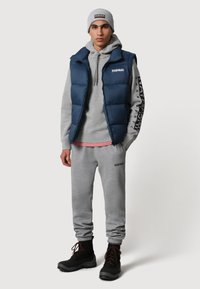 Gilet imbottito blu marina sopra una felpa grigia con cappuccio e una maglietta rosa sotto, abbinato a pantaloni della tuta grigi e stivali neri con lacci. Berretto grigio con dettaglio del logo.