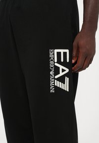 Pantalones atléticos negros con un logo bordado en blanco, que incluye "EMPORIO ARMANI" y "EA" en el costado, fabricados con una tela suave.