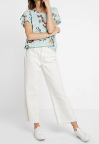 Světle modrá květinová blůza s krátkými volánkovými rukávy, spárovaná s bílými širokými krátkými džíny a bílými teniskami. Jednoduché pozadí.