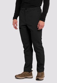 Sorte softshell-bukser har en afslappet pasform, elastisk linning, sidelommer og forstærkede knæ. De er parret med brune vandrestøvler.