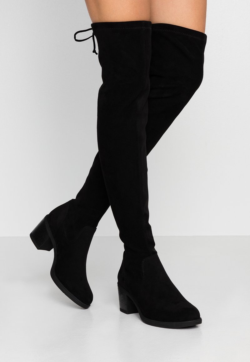 Femme portant des cuissardes en daim noir avec talons blocs, debout sur un sol blanc réfléchissant devant un fond gris uni.