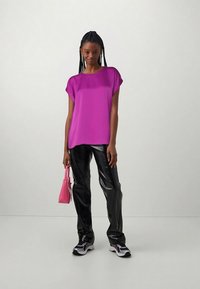 Γυναίκα που στέκεται φορώντας φωτεινό μωβ μπλουζάκι, μαύρο γυαλιστερό παντελόνι, αθλητικά παπούτσια και κρατώντας μία μικρή ροζ τσάντα ενάντια σε ένα απλό φωτεινό φόντο.