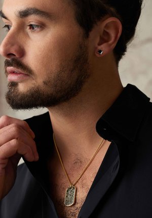 Gouden ketting met een sierlijke zwarte dogtag hanger. Model draagt een donkere shirt, waardoor de ketting en oorbel goed zichtbaar zijn. Gestructureerde huid zichtbaar.