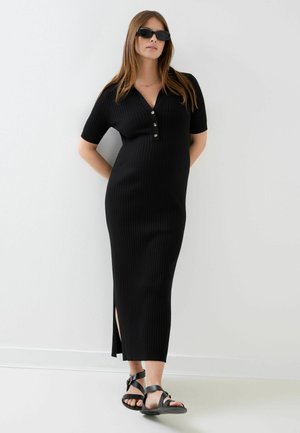 Femme portant une robe midi en maille côtelée noire avec des boutons sur le devant, une fente latérale, des sandales noires et des lunettes de soleil noires, debout contre un mur blanc.