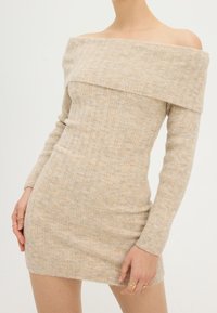 Robe beige en maille côtelée à épaules dénudées avec manches longues, présentant une silhouette ajustée et une texture douce. La longueur de la robe atteint le milieu des cuisses.