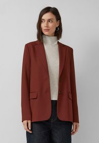 Vrouw in een roestrode blazer over een lichtgrijze coltrui en donkere jeans, die naar links kijkt tegen een effen grijze achtergrond.