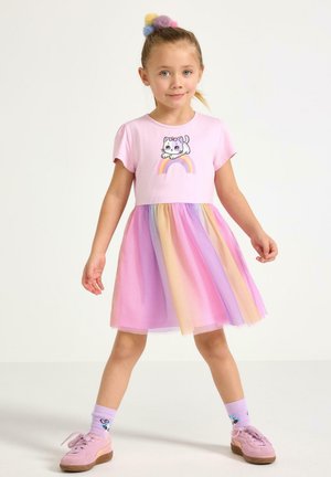 Mladá dívka oblečená v pastelově růžových šatech s obrázkem kreslené kočky a duhy, laděné sukni s duhovým motivem, pastelových ponožkách a růžových teniskách, pózující sebevědomě.