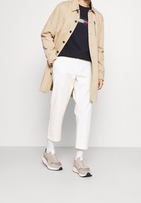 Trench coat beige, felpa blu navy con logo, pantaloni bianchi a vita alta, sneakers beige e grigie. Tessuto liscio, dettagli con bottoni, vestibilità casual.