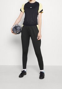 Schwarzes Sportshirt mit gelben Akzenten und Logo, kombiniert mit schwarzen Hosen, hält einen schwarzen Fußball mit weißem Text und trägt schwarze Schuhe.