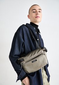 Szürke crossbody táska textúrált anyagból, zipzáros zsebbel és állítható pánttal. A modellen farmering és garbó van.