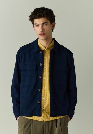 Young man wearing a navy textured buttoned jacket over a mustard yellow patterned shirt and olive green pants, standing against a plain background.