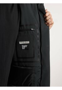 O vestimenta exterioară neagră, dotată cu o buzunar cu fermoar și un eticheta în contrast. Captuseala interioară este netedă, cu cusături vizibile și nasturi.