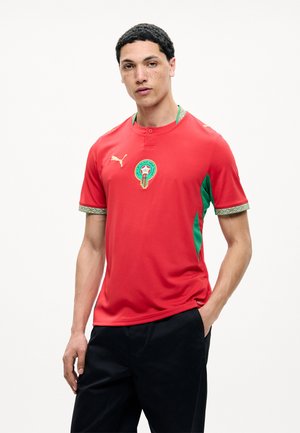 Jonge man die een rood Puma Marokko voetbalshirt met groene accenten draagt, staand tegen een effen witte achtergrond.