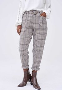 Femme portant un pantalon à carreaux gris taille haute avec ceinture et ourlets retroussés, associé à des bottines en cuir marron et un haut blanc à manches longues.