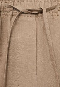 Tessuto di lino beige con una sottile texture; presenta un elastico in vita con un cordoncino annodato e dettagli cuciti lungo i lati.
