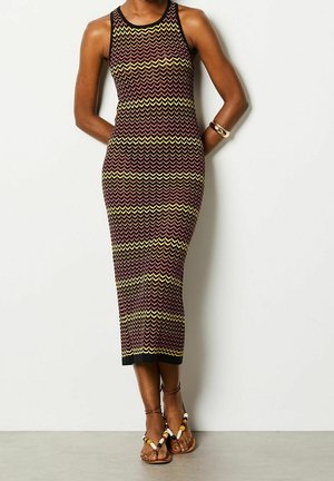 Vestito midi senza maniche, aderente, con motivo a zigzag in nero, giallo e rosso, indossato con sandali bassi con perline e braccialetti dorati.