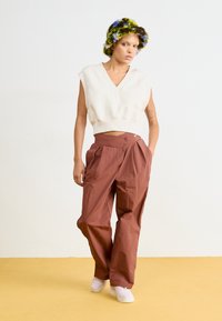 Maglione senza maniche color beige chiaro, design a scollo a V; pantaloni a gamba larga color ruggine con pieghe; sneaker pastello; berretto in maglia multicolore.